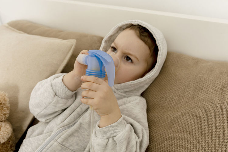 Sick little boy with inhaler for cough treatment. unwell kid doing inhalation on his bed. flu season