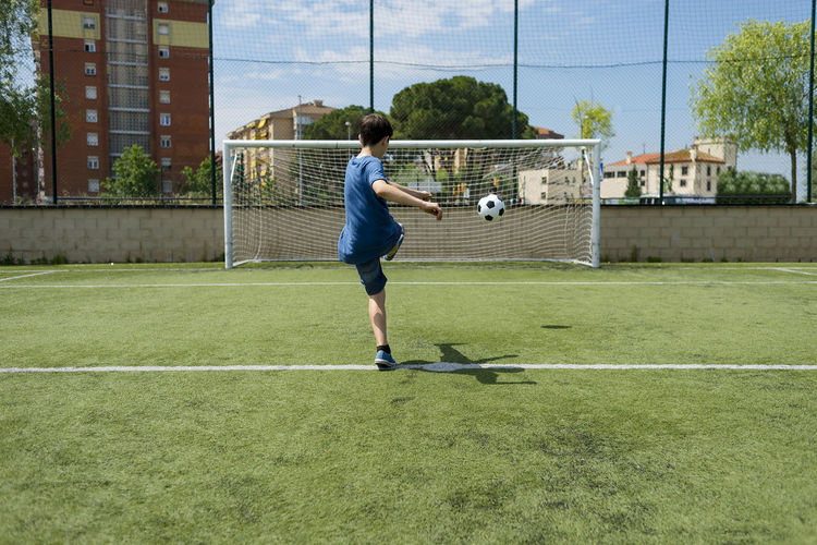 Rear view of boy kicking soccer ball towards net on field