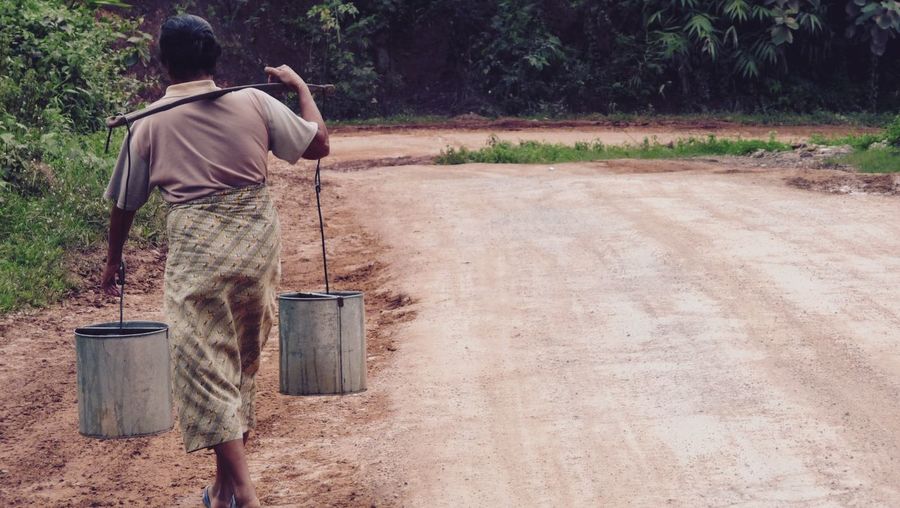 Rear view of man carrying containers on shoulder at roadside