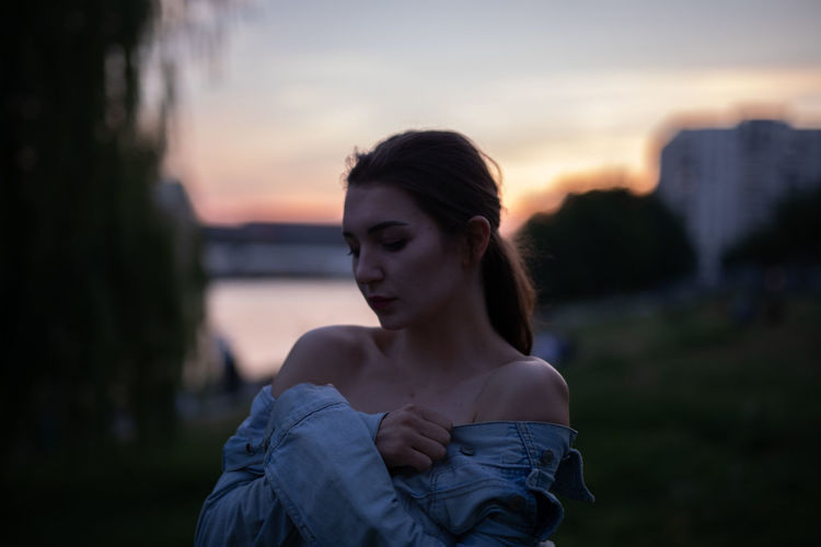 Young woman looking away while standing outdoors during sunset