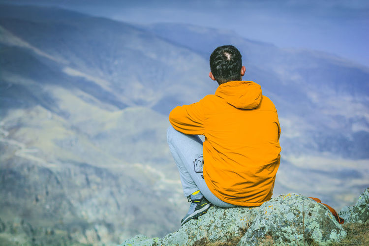 Rear view of man sitting on cliff against mountains
