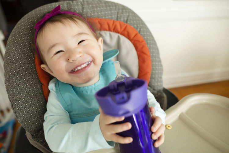 Smiling baby girl holds cup of milk while sitting in highchair at home