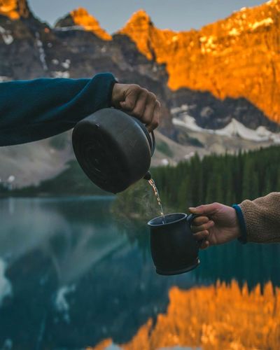 Cropped hands of person pouring drink to friend against lake