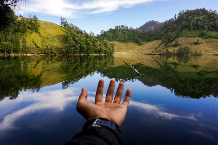 Cropped hand of man gesturing by lake against mountains
