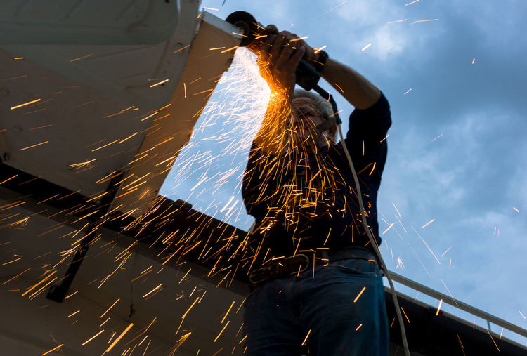 Low angle view of senior worker cutting metal against cloudy sky
