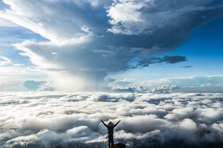 Rear view of excited man standing on mountain by clouds against sky