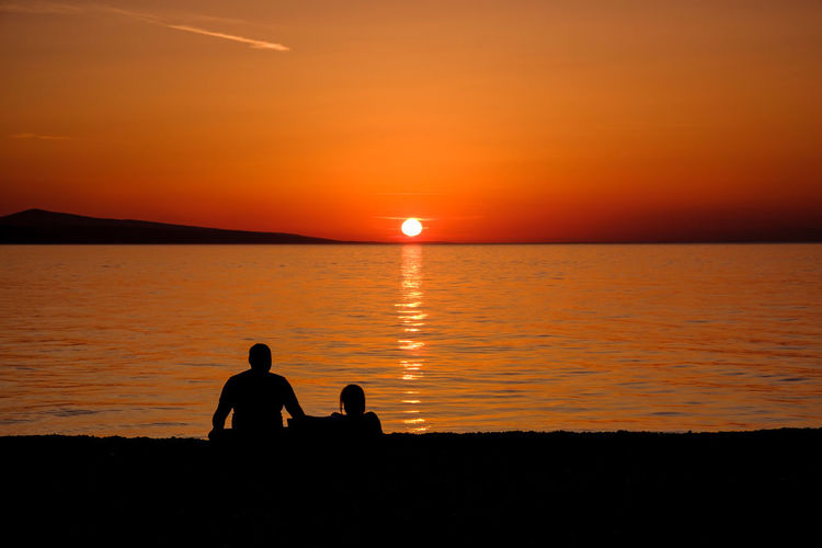Silhouette couple sitting on beach against orange sky
