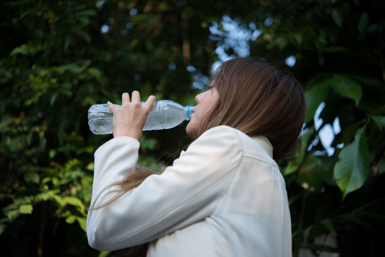 Young businesswoman drinking water from bottle against trees