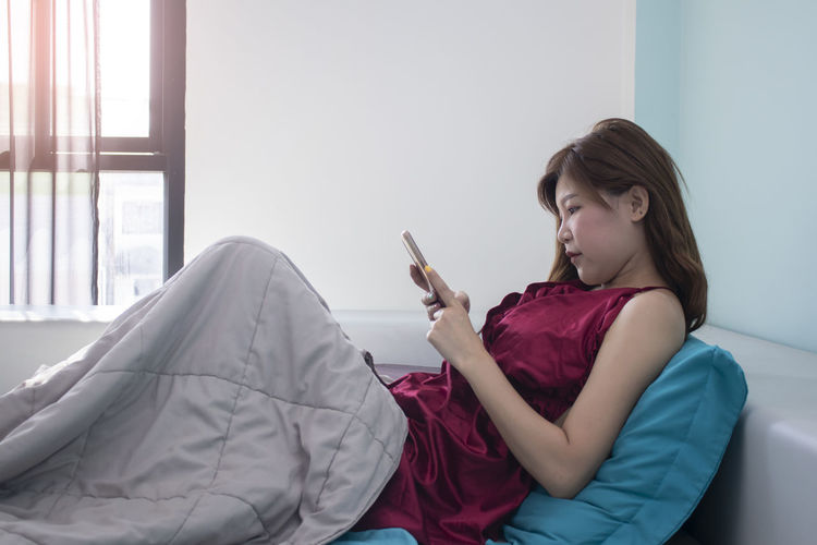 Side view of woman holding mobile phone while sitting on bed