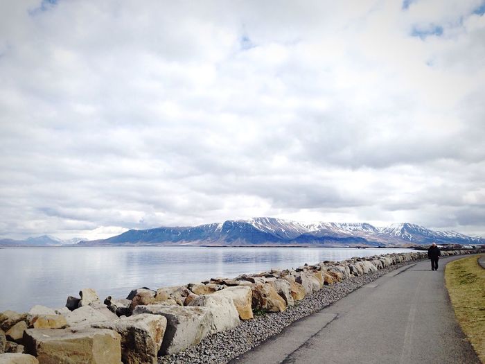 Man walking on road by lake against cloudy sky