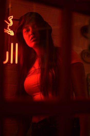 PORTRAIT OF YOUNG WOMAN STANDING AGAINST ILLUMINATED RED LIGHT