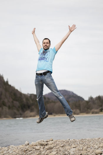 Full length portrait of man jumping against river and sky