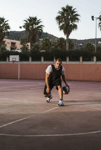 Serious determined basketball player in black and white sportswear practicing dribbling and defense tricks while training alone on sports ground against residential buildings among palms at foothill in daytime