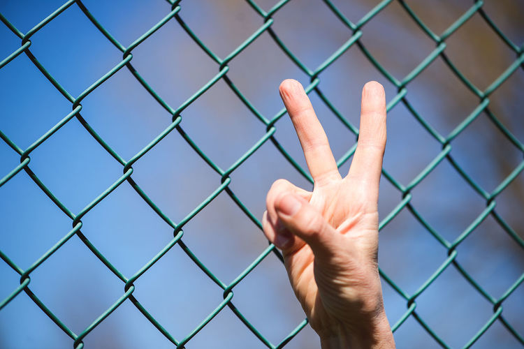 Cropped hand gesturing peace sign by green chainlink fence
