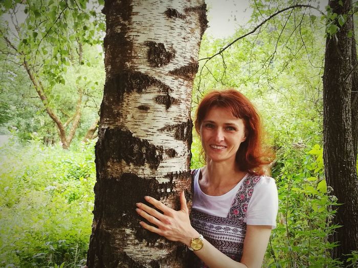 Portrait of smiling woman standing by tree trunk in forest