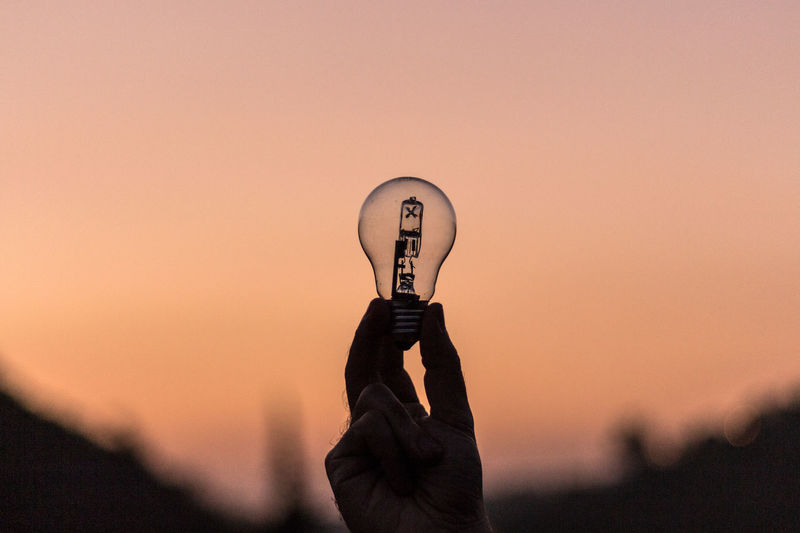 Silhouette hand holding light bulb against sky during sunset