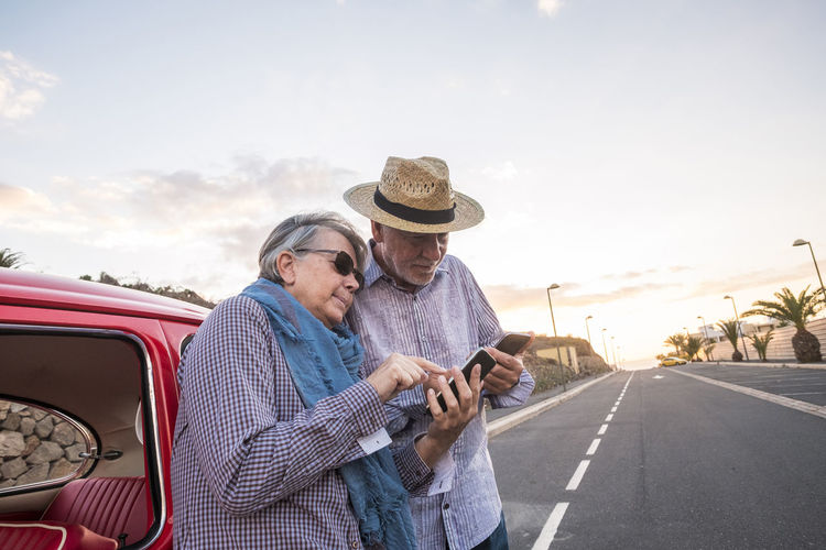 Senior couple using smart phone while standing on road against sky
