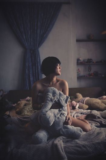 Naked woman sitting with stuffed toy on bed at home