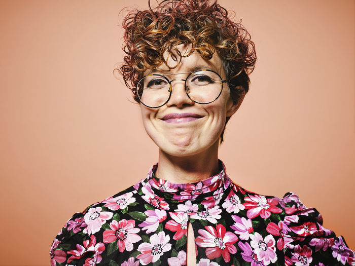 Happy young curly haired female in stylish floral blouse and trendy eyeglasses smiling and looking at camera against red background