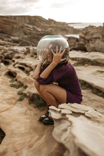Side view of female in glass astronaut helmet sitting on rocky ground and looking away while pretending being spaceman