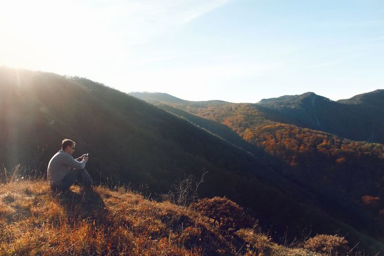 Man using mobile phone sitting on mountain against sky