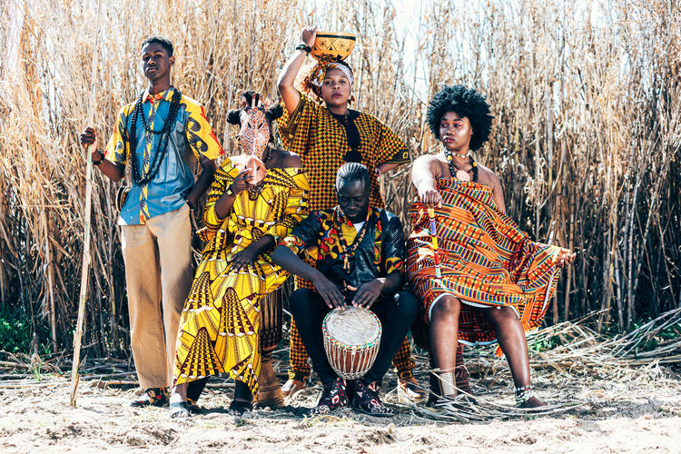 Full body group of black and mixed race women and men in colorful authentic african outfits with sticks and drums sitting and standing against dry field grass