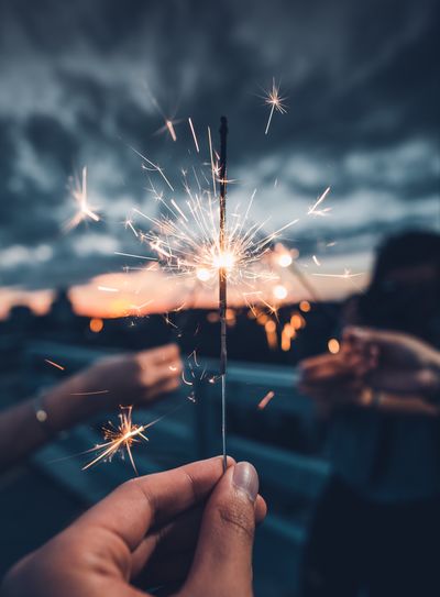 Cropped hands holding illuminated sparklers against sky during sunset