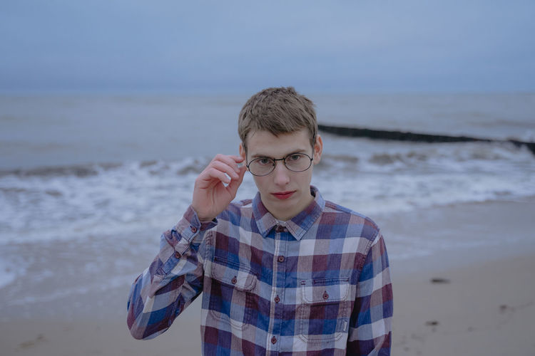 A guy in a checkered shirt, in round glasses for sight, looks at the cameras, stands on the seashore