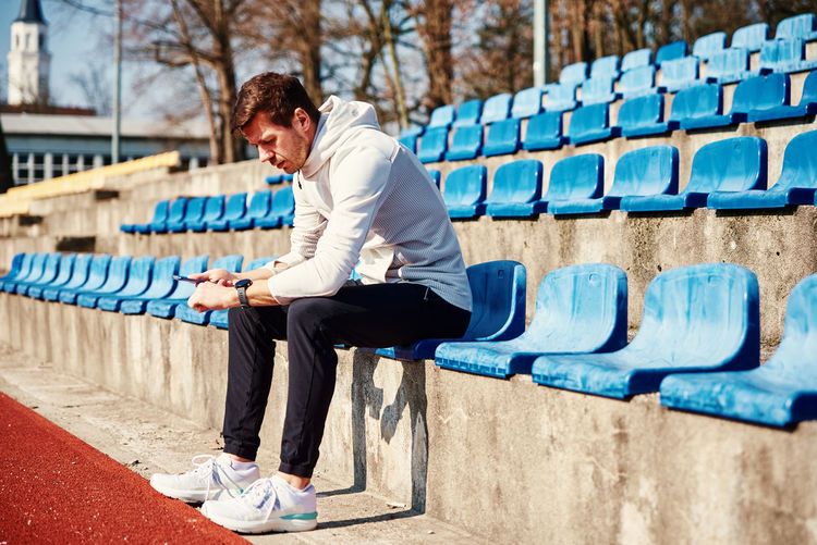 Man in sportswear sitting on stadium seat and browsing smartphone, check fitness watch