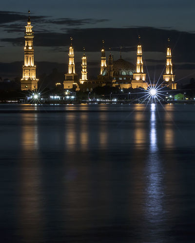 Illuminated mosque by river against sky at night