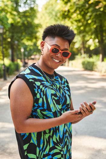 Happy filipino transgender woman in stylish clothes and sunglasses browsing cellphone and looking at camera with smile while standing on sunlit path in summer in park