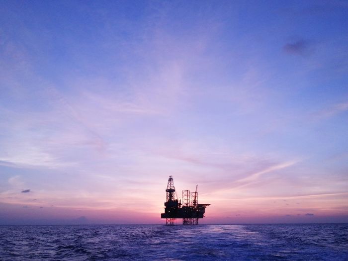 Silhouette oil rig in sea against sky during sunset