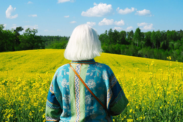 Rear view of senior woman standing on rapeseed field against sky