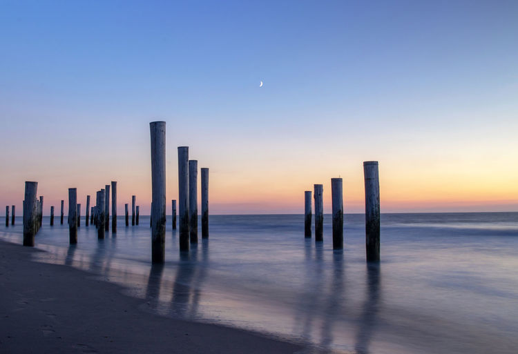 Wooden posts in sea against clear sky at sunset