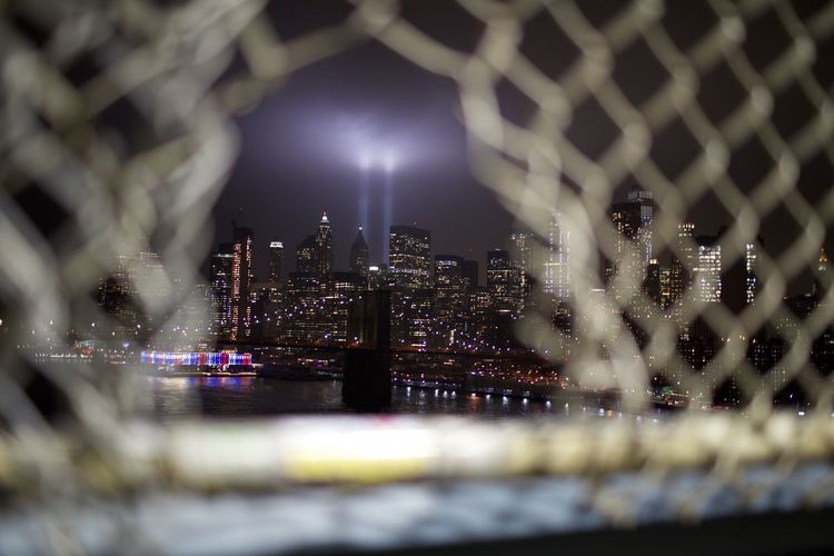 Illuminated buildings in city seen through broken chainlink fence at night