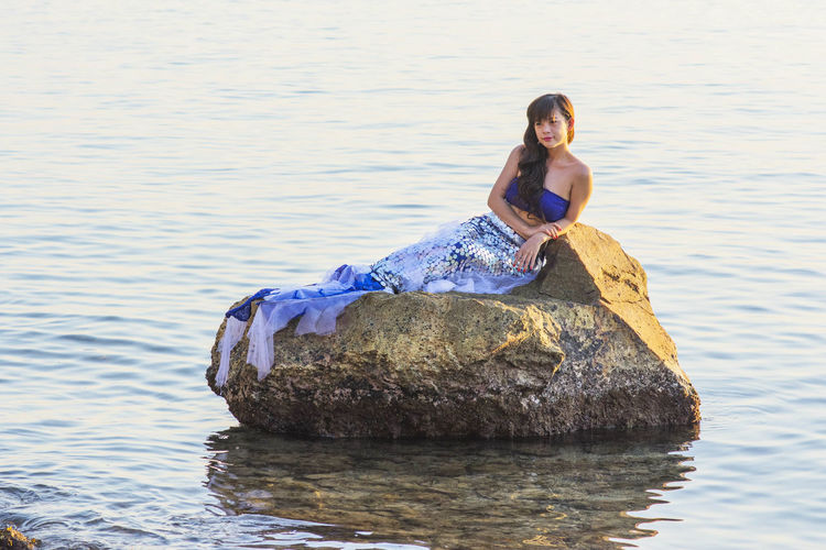 Woman in mermaid costume sitting on rock in lake