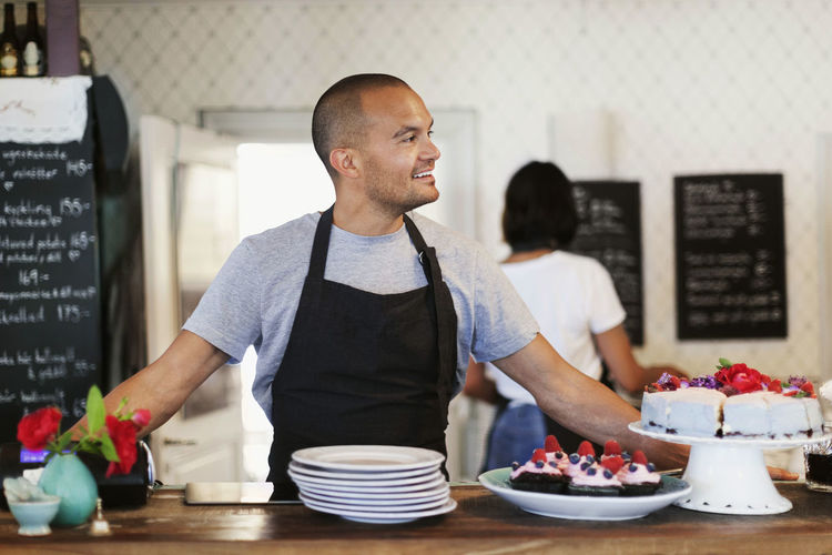 Smiling male owner standing at cafe counter with coworker working in background