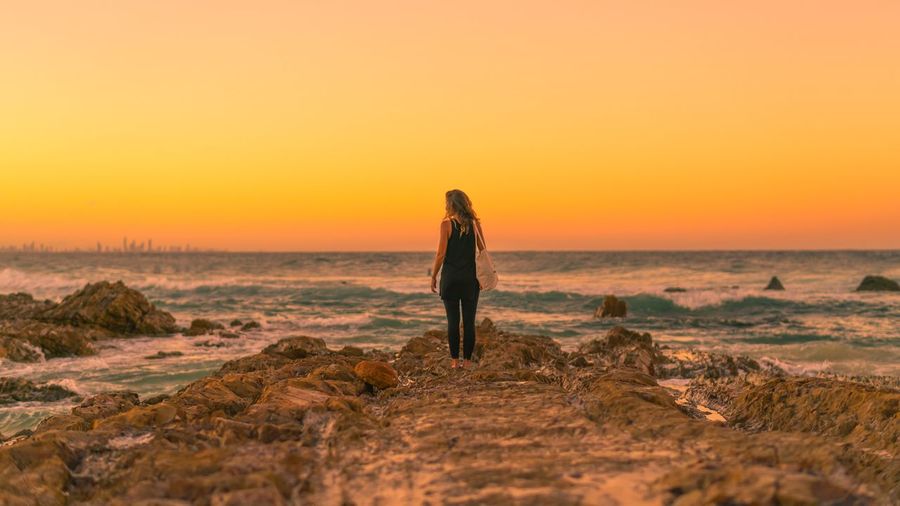 Full length rear view of woman standing at beach against clear sky during sunset
