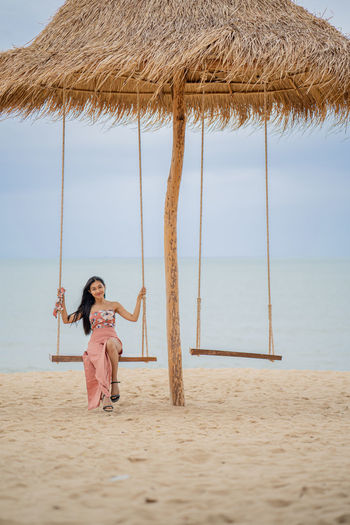Full length of woman sitting on rope swing at beach against sky
