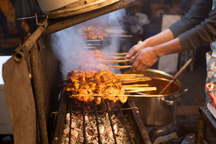 Street food cook grills chicken bbq skewers with apparent smoke