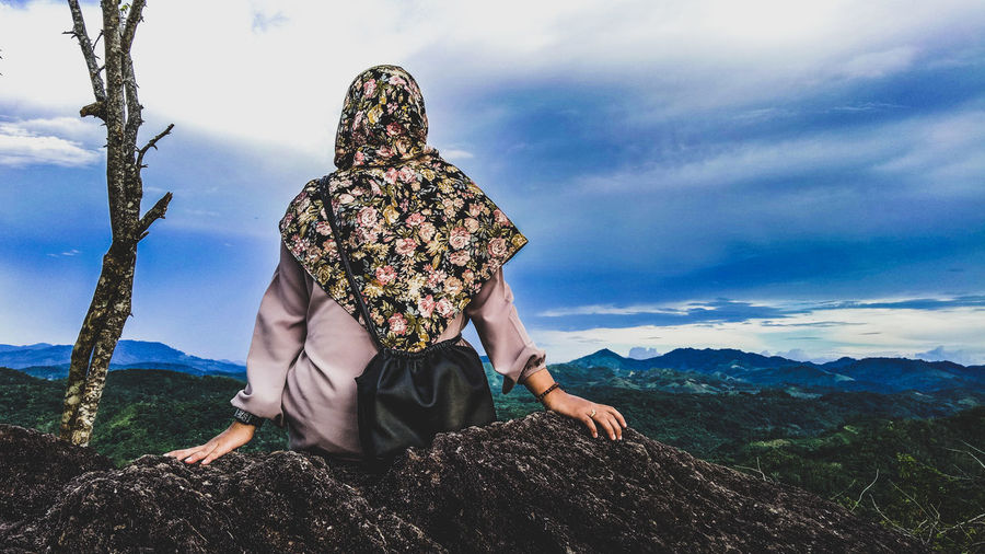 Rear view of woman sitting on cliff against cloudy sky