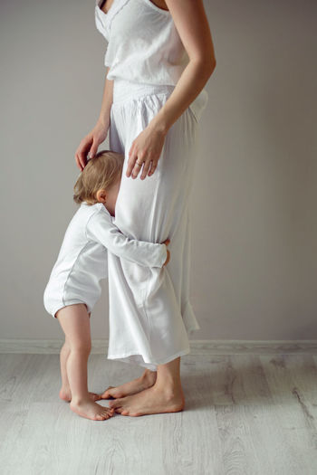Girl child stands next to her mother at the window in white clothes at home in winter
