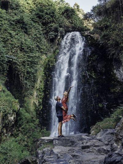 Side view of man carrying woman while standing against waterfall
