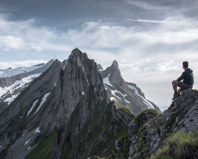 Side view of man sitting on rocky mountain against cloudy sky
