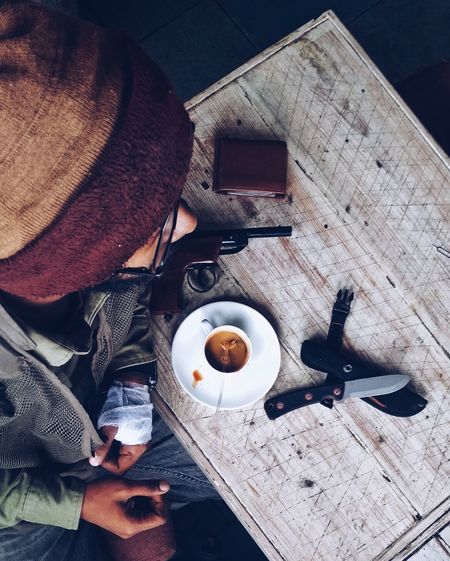 High angle view of young man sitting by coffee with knife and gun in table