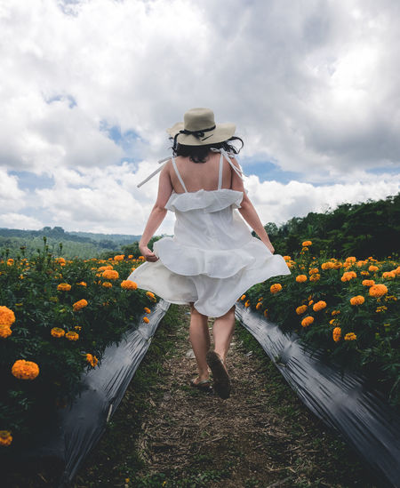 Rear view of woman walking amidst flowering plants against cloudy sky