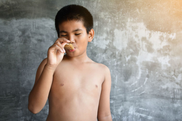Shirtless boy eating popsicle while standing against wall
