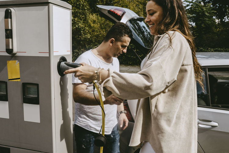 Smiling mother holding electric plug standing by father at charging station