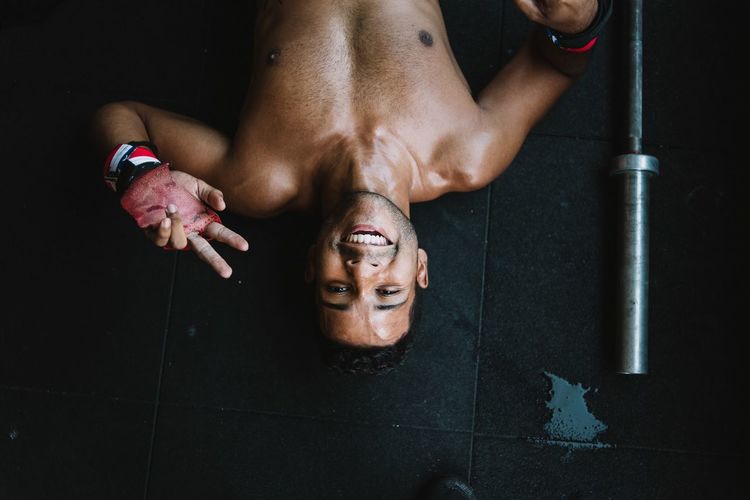 High angle portrait of shirtless man gesturing peace sign while lying on floor