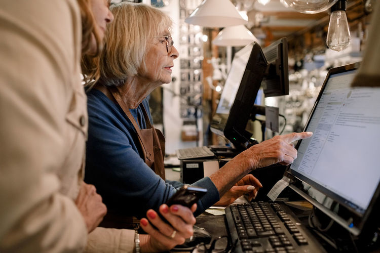 Senior female customer and saleswoman discussing over computer monitor at checkout in hardware store